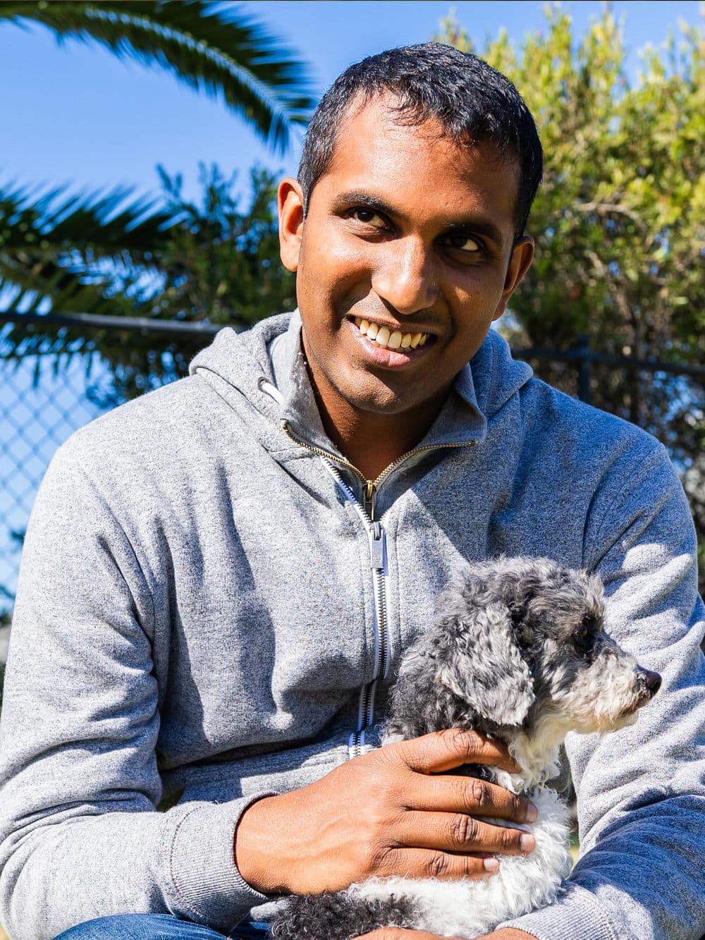 A man with short black hair and a gray hoodie is smiling and looking off to the left side while holding a small, fluffy gray and white dog in his arms. The dog is looking to the right.