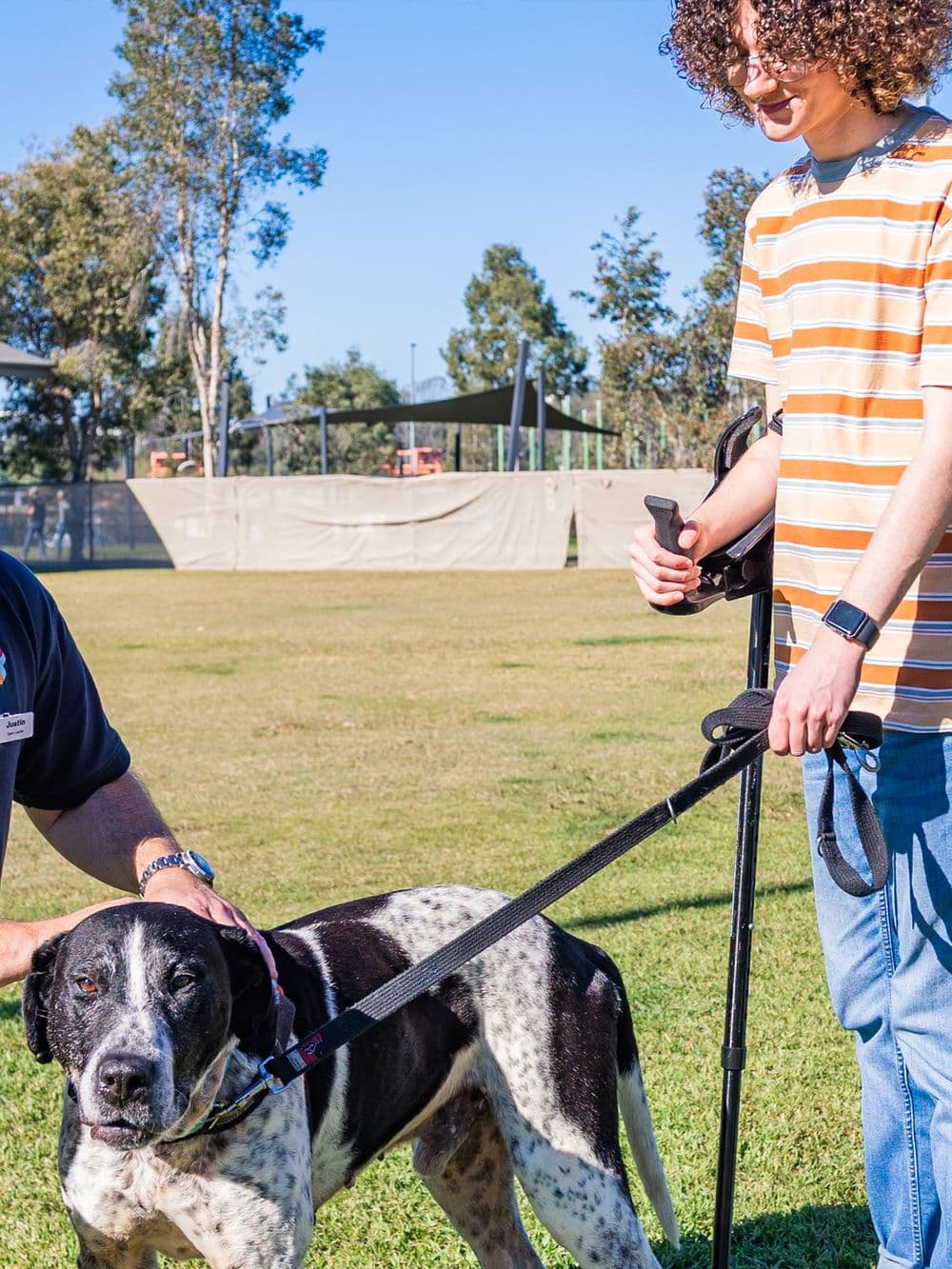 Outdoors on a grassy field, a man identified as Trainer Justin, and a young man with curly hair, identified as Alex, are with a large black and white speckled dog. Justin is kneeling and petting the dog's head, while Alex, holding a walking stick and the dog's leash, is smiling down at the dog.