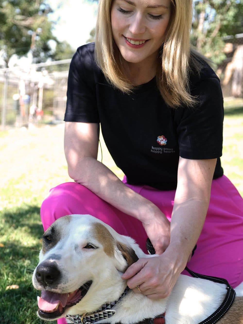 A young woman with blonde hair, identified as Zoe, kneels on a grassy area outdoors. She is wearing a black t-shirt with a logo and bright pink pants. She is smiling down at a large white and brown rescue dog and is gently petting it under the chin. The dog is looking up with a happy, panting expression.